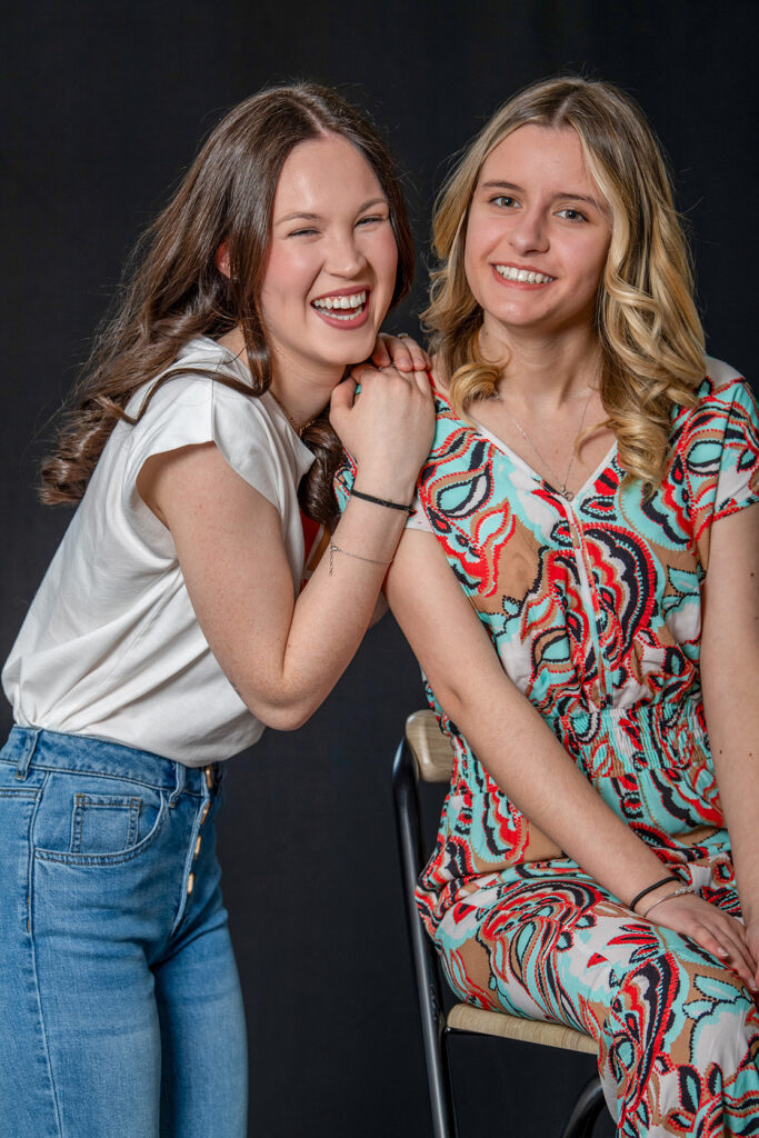 Deux jeunes femmes souriantes posant ensemble, l’une debout en jean et t-shirt blanc, l’autre assise en combinaison colorée, sur fond noir. Photo de Dominique Ducasse. Photographe à Poitiers.